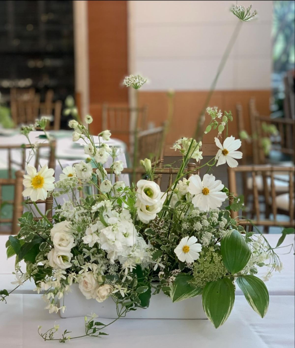 A low white centerpiece of garden roses, cosmos, lisianthus and greenery on a reception table