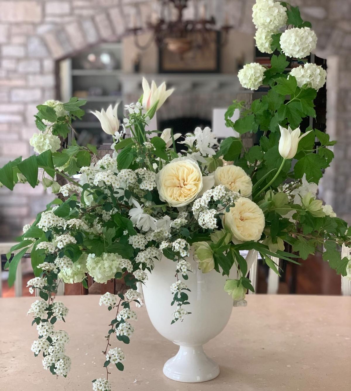 A tall white urn arrangement of snowball viburnum, spirea, garden roses and white tulips
