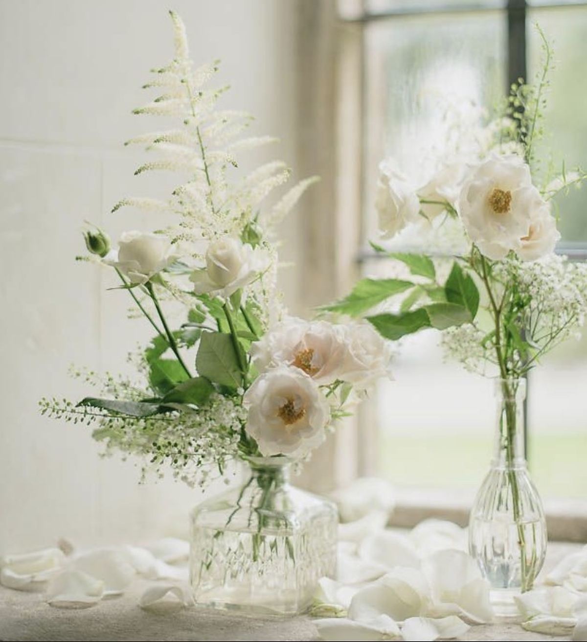 White garden roses, astilbe and baby&rsquo;s breath in two clear glass bud vases by a window