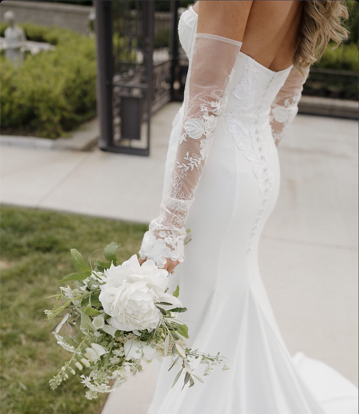 A bride in a lace-sleeved gown holding a loose white bouquet of peony, foxglove and garden greenery